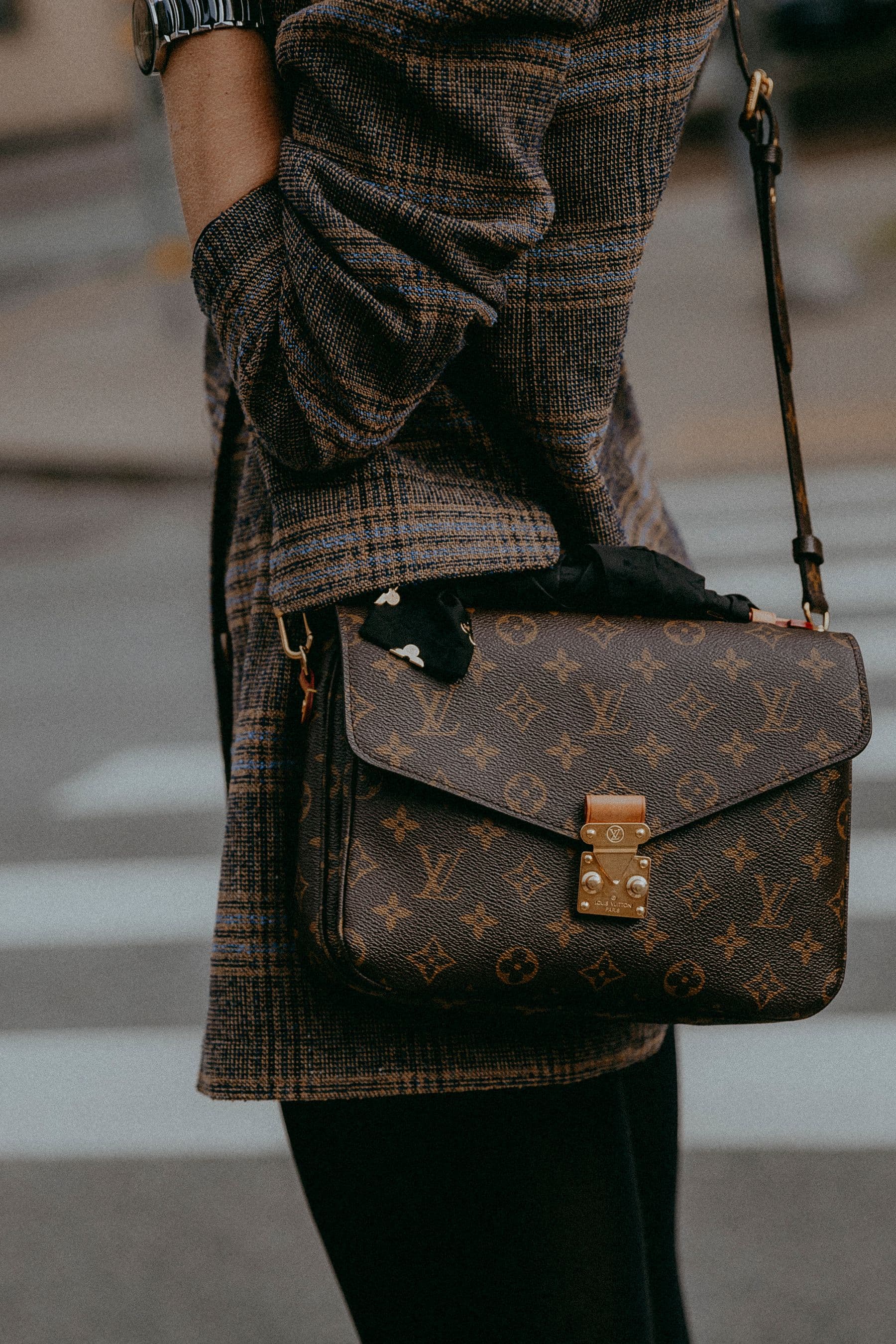A Louis Vuitton monogram crossbody carried with a plaid coat on a city street